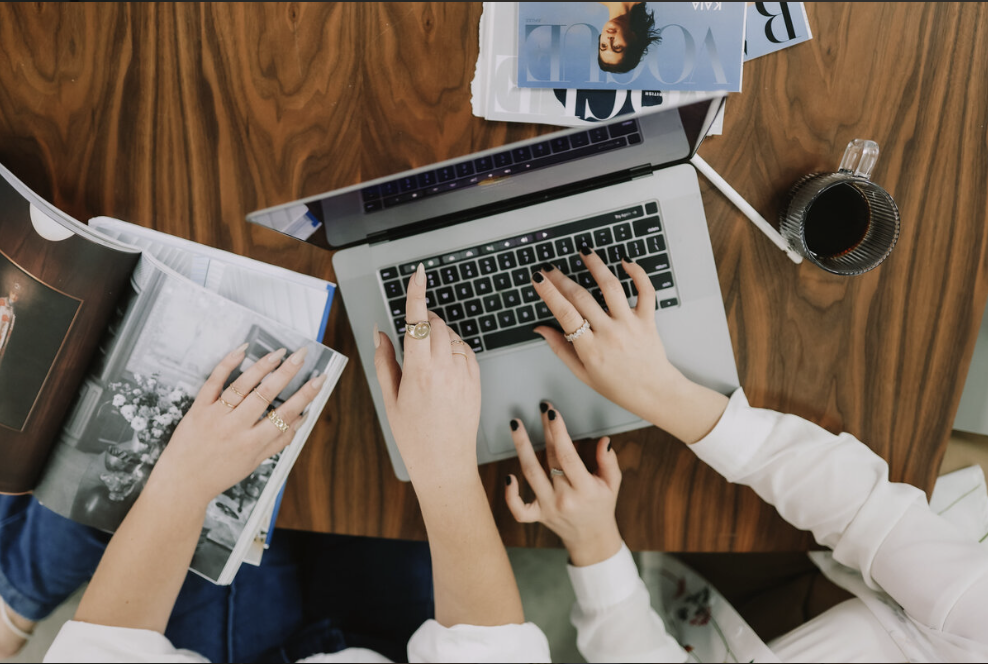 A laptop computer on a desk surrounded by magazines and other work and two females arms pointing at the screen.